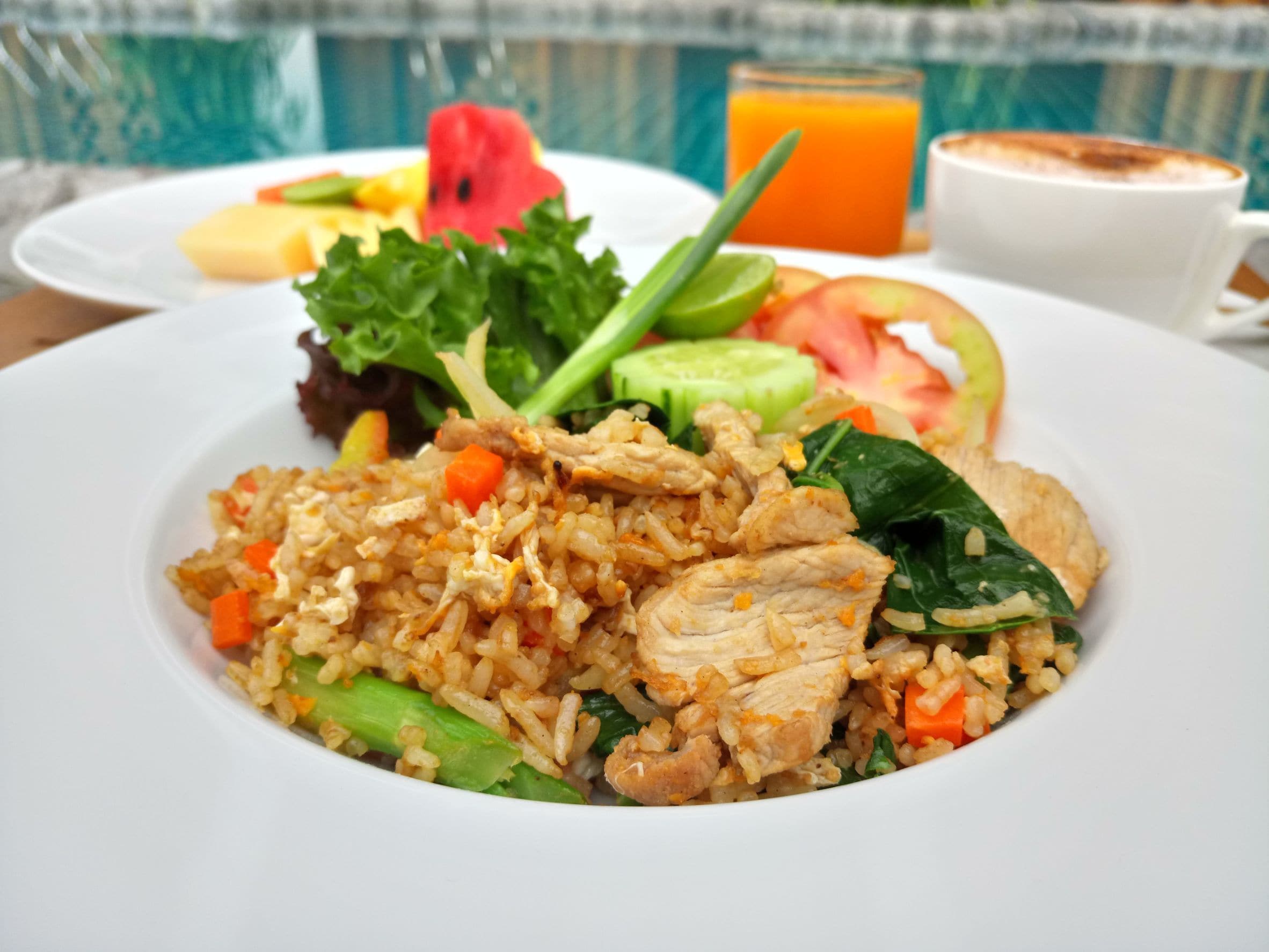 Une assiette de riz frit aux légumes et au poulet, un verre de jus d’orange et une tasse de café sur une table.