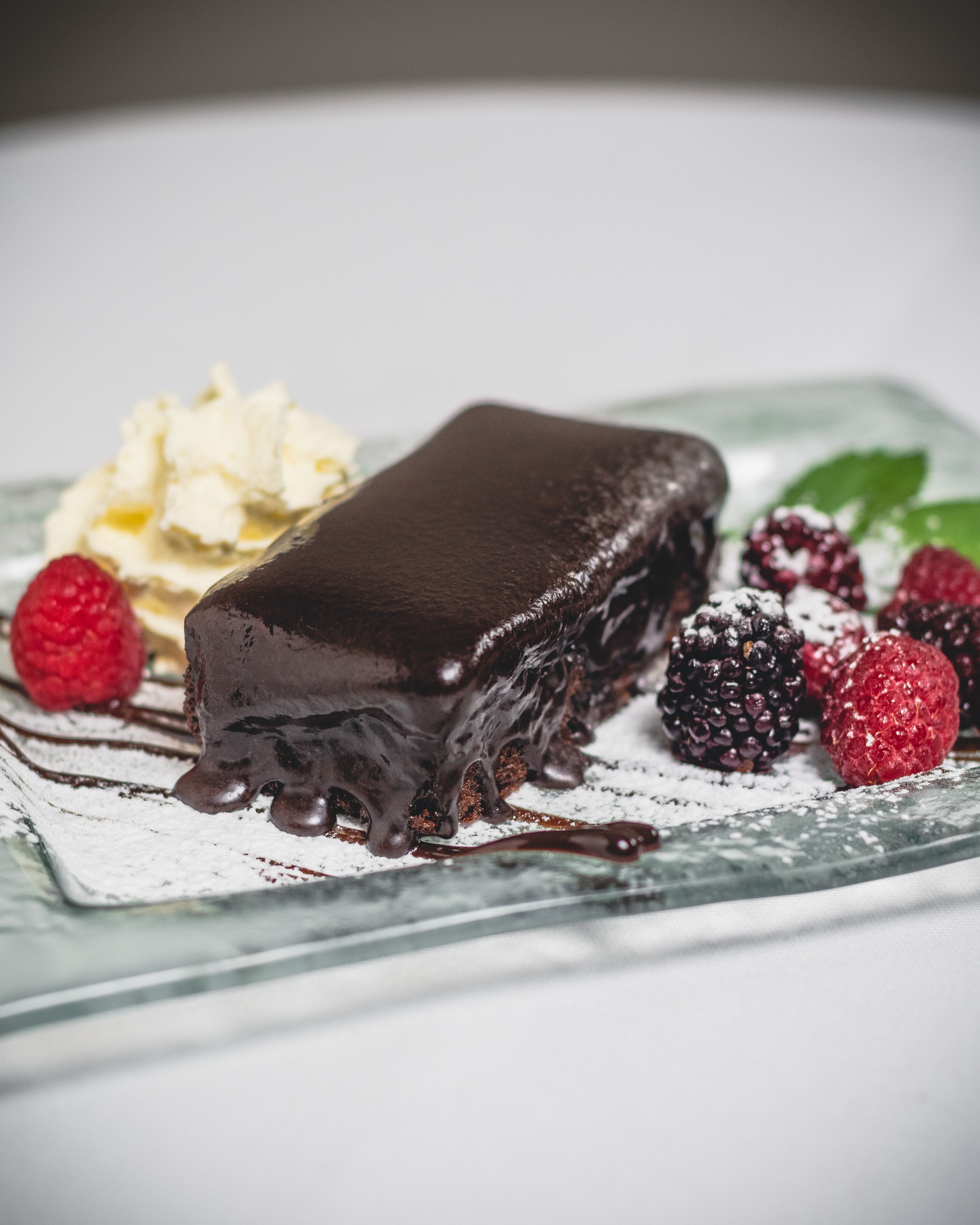 Un postre con glaseado de chocolate, frambuesas y crema batida sobre un plato de cristal.