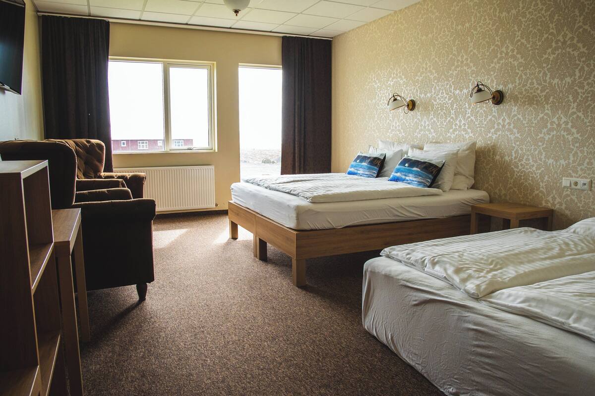 A hotel room with a bed, a desk, and a chair. There is a window with curtains, a radiator, and a wall with patterned wallpaper.