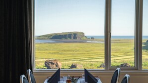 A dining table with chairs and a view of a coastal landscape through large windows. - Hótel Dyrhólaey (Vik I Myrdal)