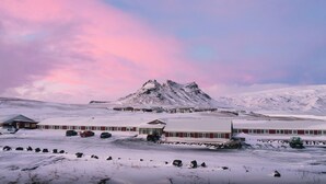 A snow-covered landscape with a mountain in the background, a building with a red facade, and a clear sky. - Hótel Dyrhólaey (Vik I Myrdal)