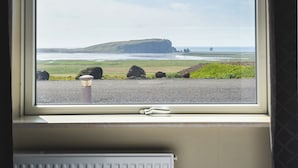 A room with a large window overlooking a coastal landscape, including a grassy area and a rocky outcrop. - Hótel Dyrhólaey (Vik I Myrdal)