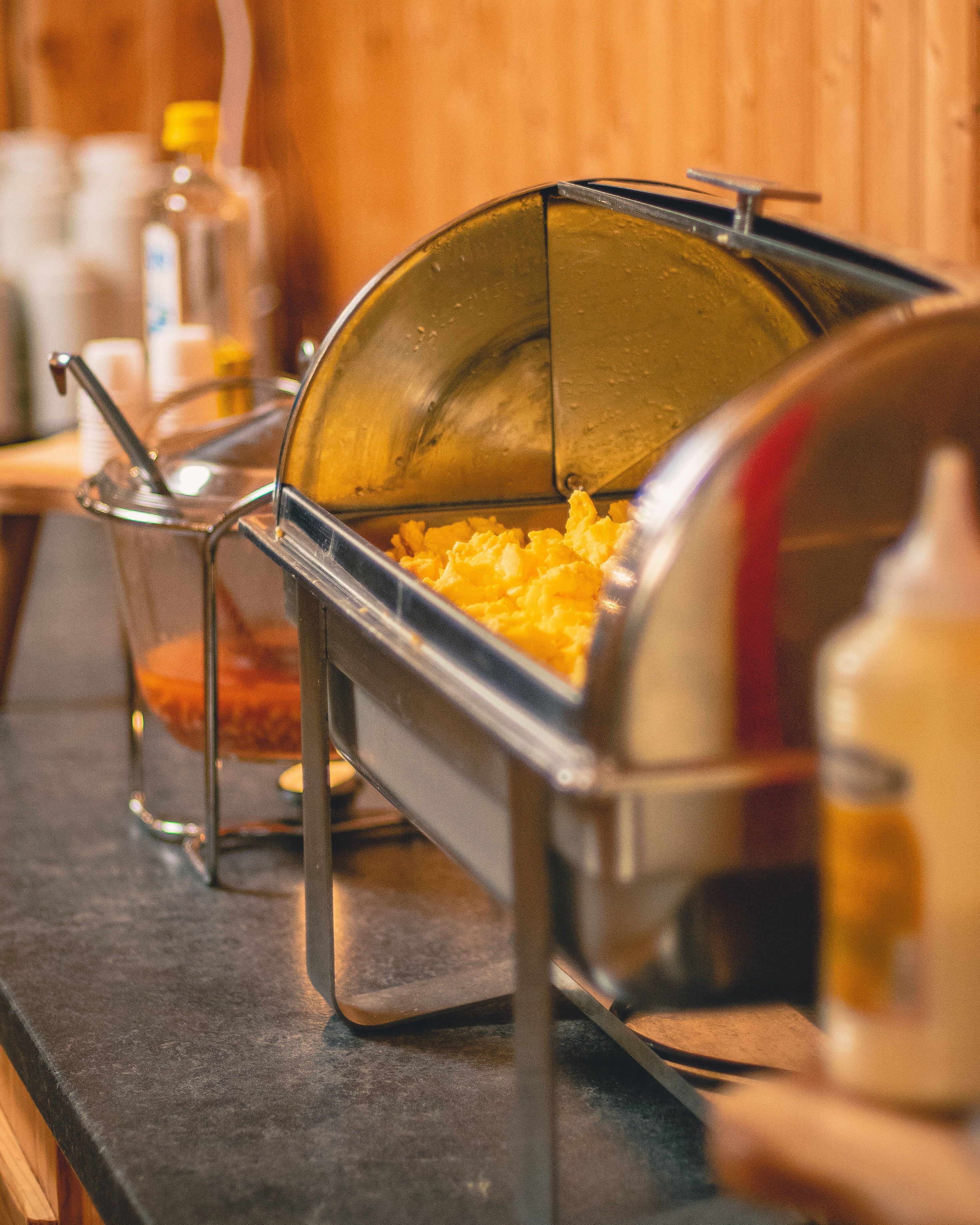 Una mesa de buffet con un recipiente cubierto que contiene huevos revueltos, un recipiente de vidrio con un líquido rojo, y una botella de aceite.