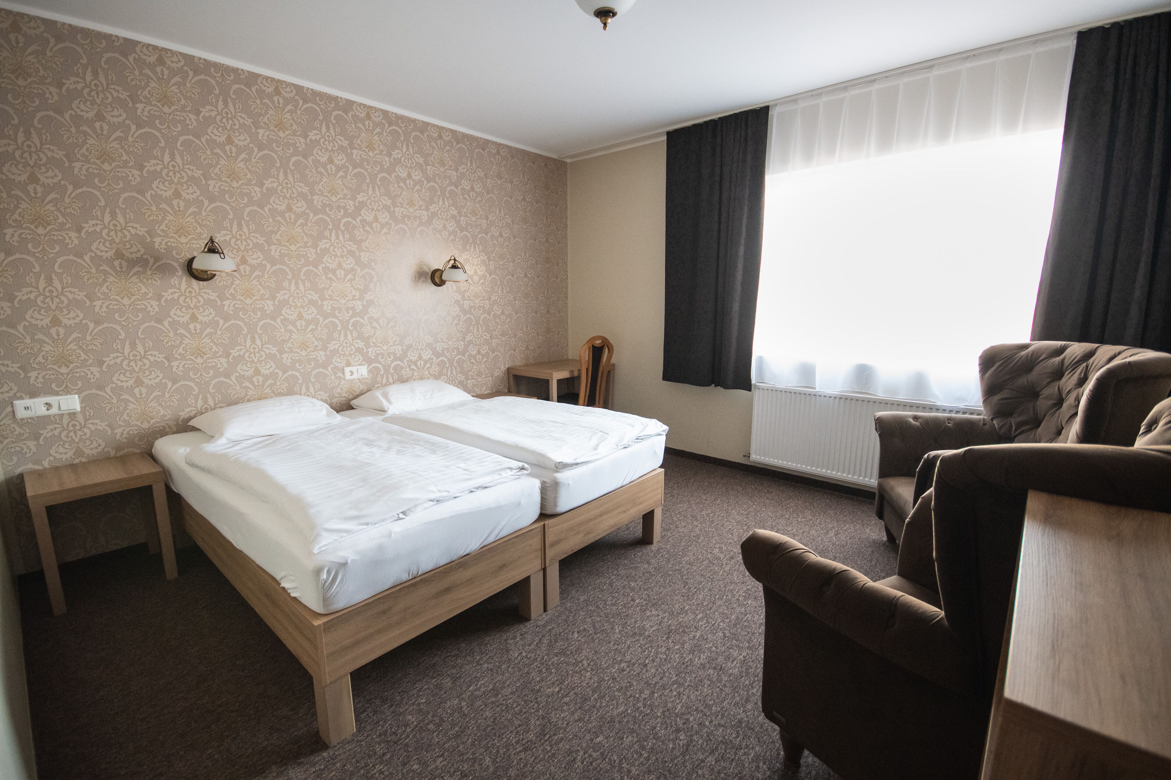 Habitación de hotel con dos camas individuales, una mesita de noche de madera, una silla y un ventanal con cortinas.