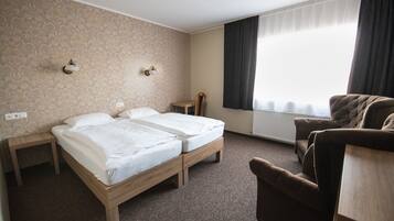 Habitación de hotel con dos camas individuales, una mesita de noche de madera, una silla y un ventanal con cortinas.