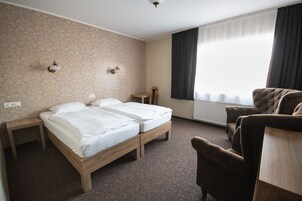 Habitación de hotel con dos camas individuales, una mesita de noche de madera, una silla y un ventanal con cortinas.