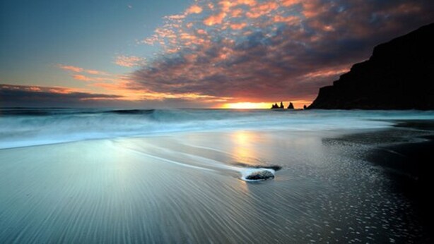A beach with a sunset, waves, and a silhouette of a cliff. - Hótel Dyrhólaey (Vik I Myrdal)