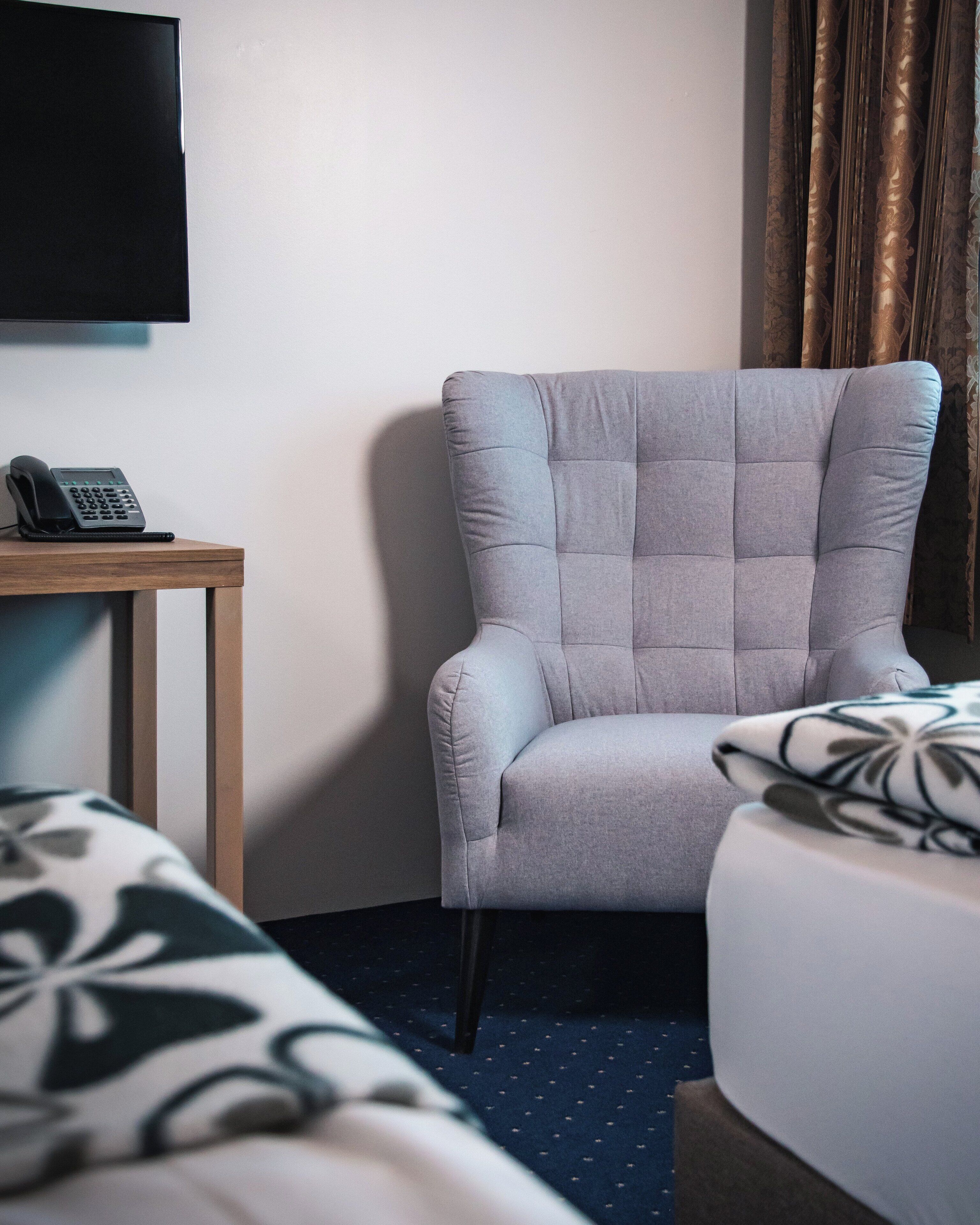 Quarto de hotel com uma poltrona estofada cinza, uma pequena mesa de madeira com um telefone e uma cama com colcha estampada.