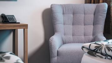 Quarto de hotel com uma poltrona estofada cinza, uma pequena mesa de madeira com um telefone e uma cama com colcha estampada.
