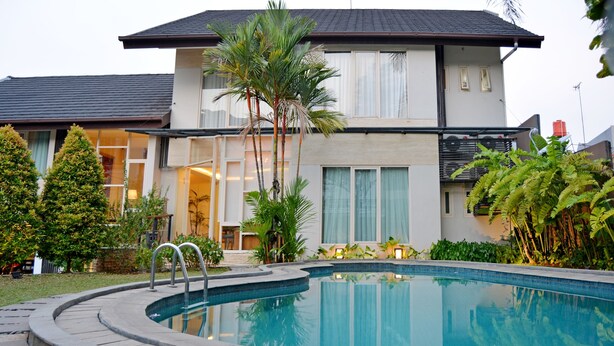 A swimming pool with a curved edge, surrounded by a stone deck. There are palm trees and other greenery around the pool area. - W Home Cipete (Jakarta)