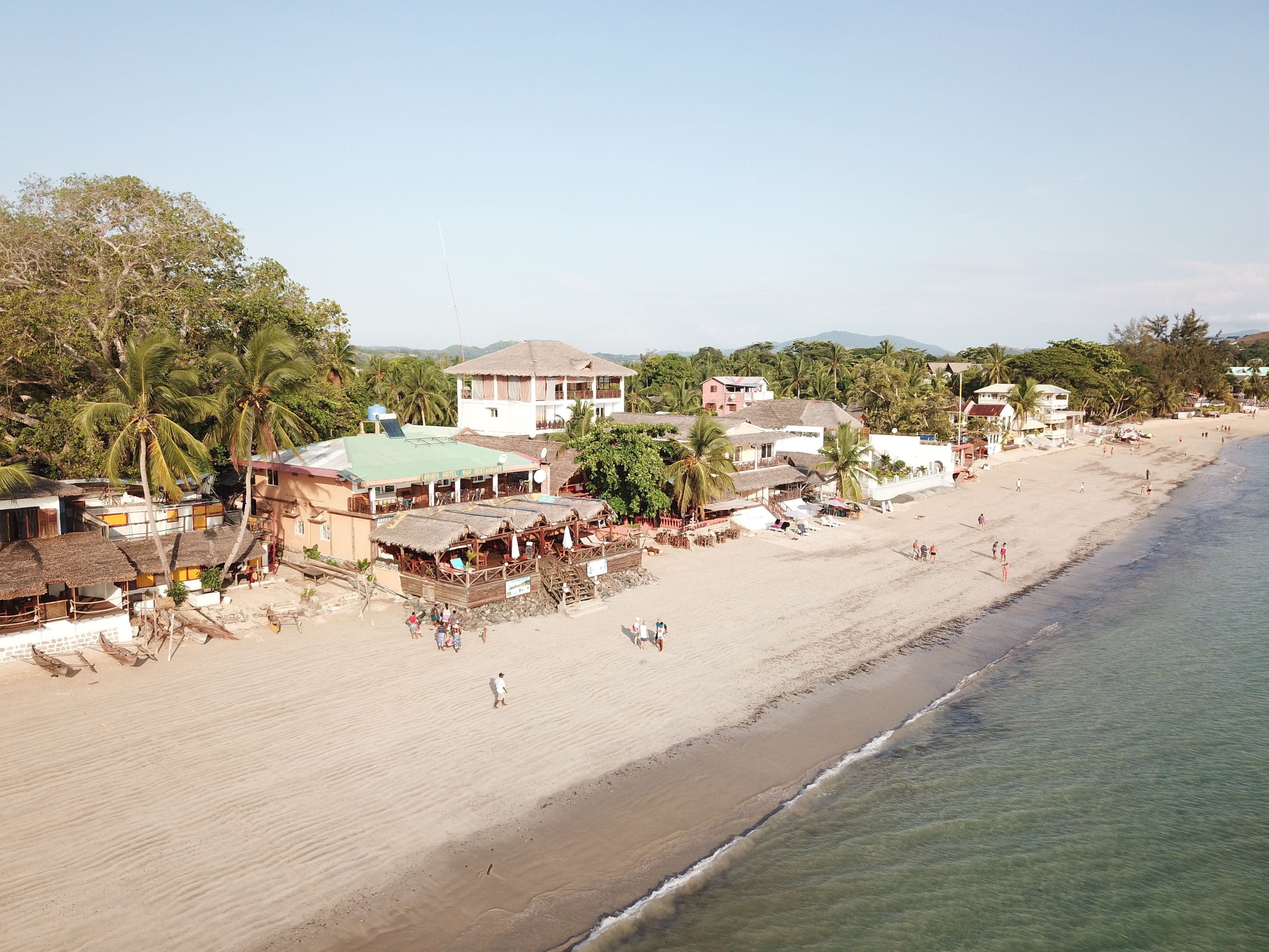 Un hotel frente a la playa con varias construcciones, que incluyen un restaurante y un bar, y una playa de arena con gente y palmeras.