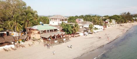 Un hotel frente a la playa con varias construcciones, que incluyen un restaurante y un bar, y una playa de arena con gente y palmeras.
