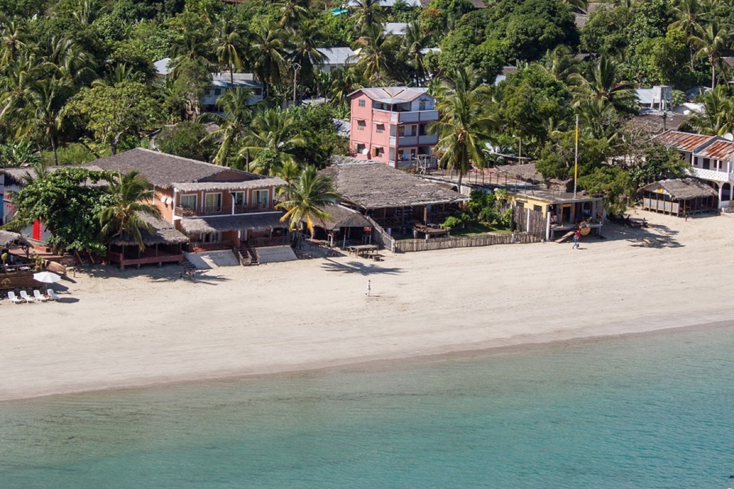 Une propriété située en bord de mer, comprenant plusieurs bâtiments, dont une imposante structure rose et plusieurs petites cabanes, le tout entouré de palmiers et bordé d’une plage de sable.
