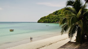 Une plage à l’eau turquoise et limpide, un rivage sablonneux et, en arrière-plan, une colline verdoyante.
