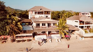Il s'agit d'une propriété située en bord de mer, comprenant plusieurs bâtiments, dont une imposante structure blanche surmontée d'un toit de chaume. On y trouve plusieurs chaises de plage et des parasols, et des palmiers sont visibles.