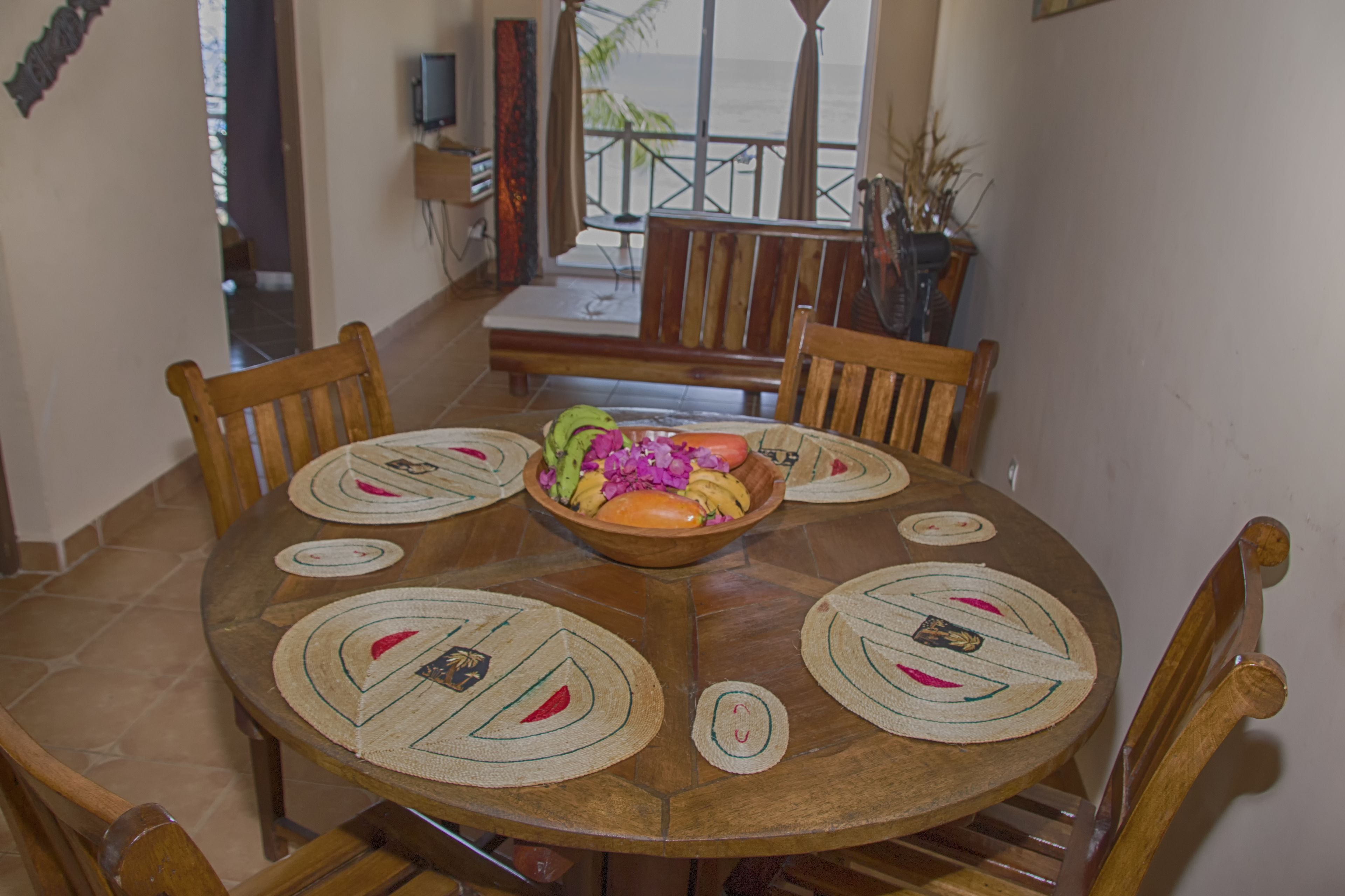 Una mesa de comedor redonda de madera con cuatro sillas, un cuenco central con frutas y flores, y posavasos con diseño de hojas.