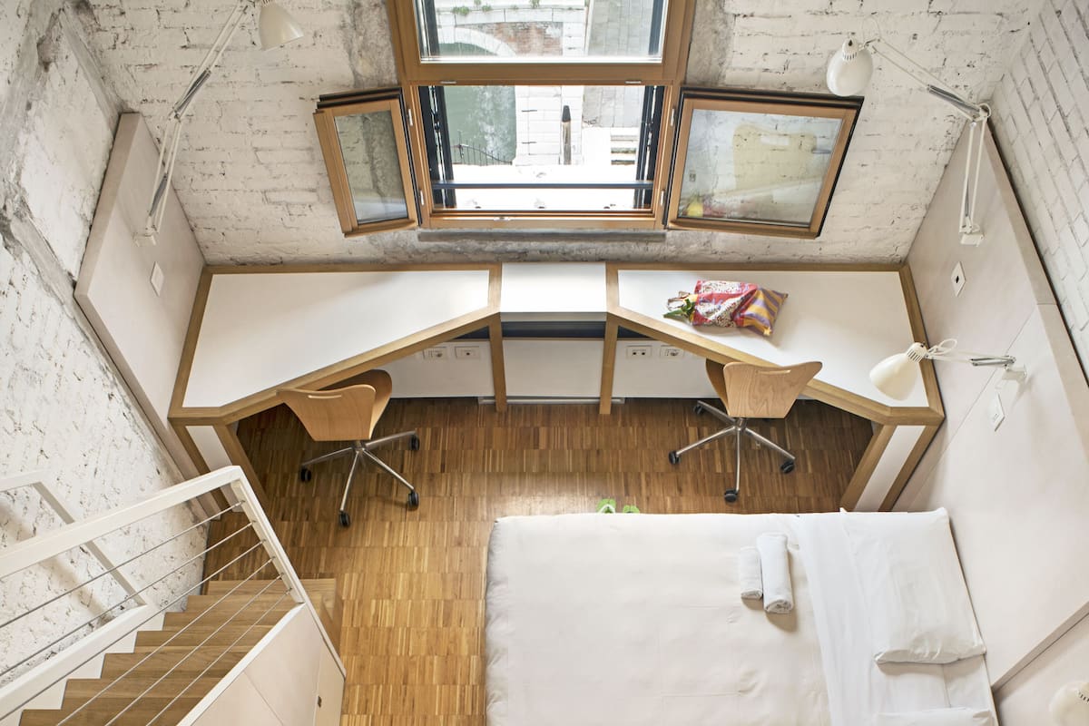 A compact room with a bed, desk, and chair, a window with a view, and a staircase.