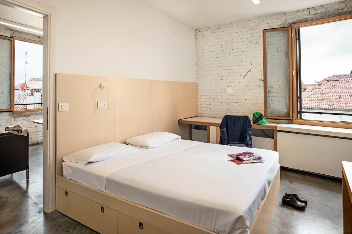 A bedroom with a bed, a desk, a chair, a window, and a bathroom.