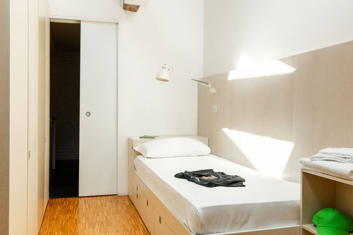 A small, neatly organized bedroom with a single bed, a nightstand, a desk, and a shelf with towels.