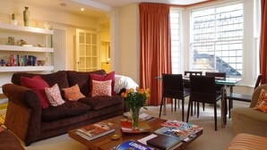 A living room with a brown sofa, a coffee table with magazines, a vase with flowers, and a dining area with chairs and a table. - The London Agent Chelsea Finborough (London)