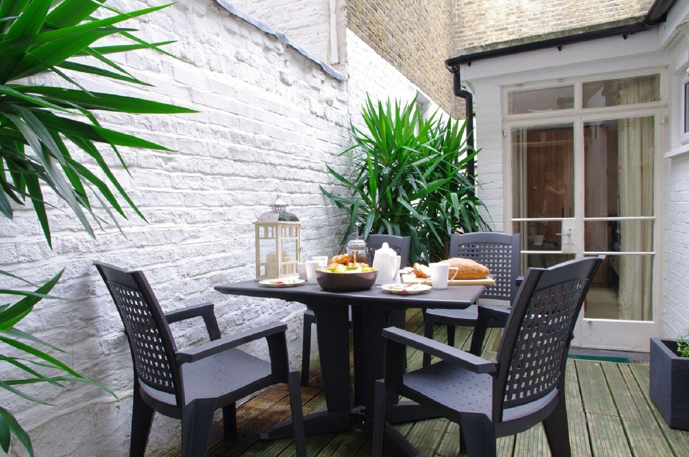 A patio with a table set for two, surrounded by black chairs, a white brick wall, and a large green plant.
