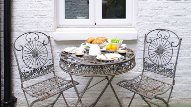 A round metal table with two chairs, a white window, and a small plant inside. - The London Agent Chelsea Finborough (London)