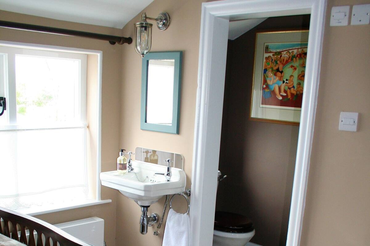 A bedroom with a bed, a bedside table, a window, a bathroom with a sink and toilet, and a door leading to another room.