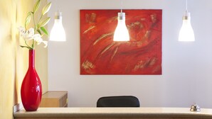 A reception desk with a red vase of flowers, two hanging light fixtures, and a red abstract painting on the wall. - Hotel Drei Morgen (Leinfelden-Echterdingen)
