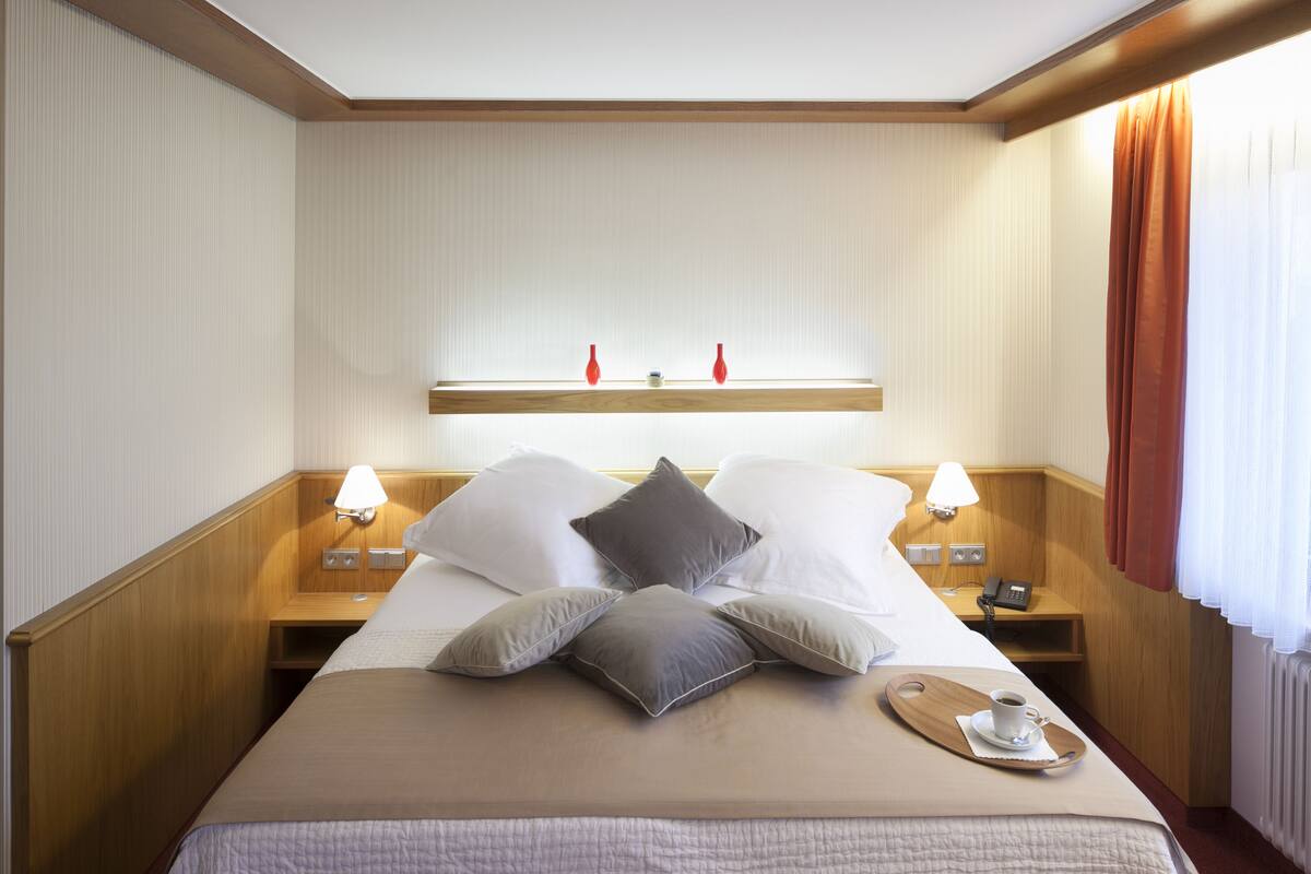 A hotel room with a neatly made bed, bedside lamps, a small table with a cup, and a window with curtains.