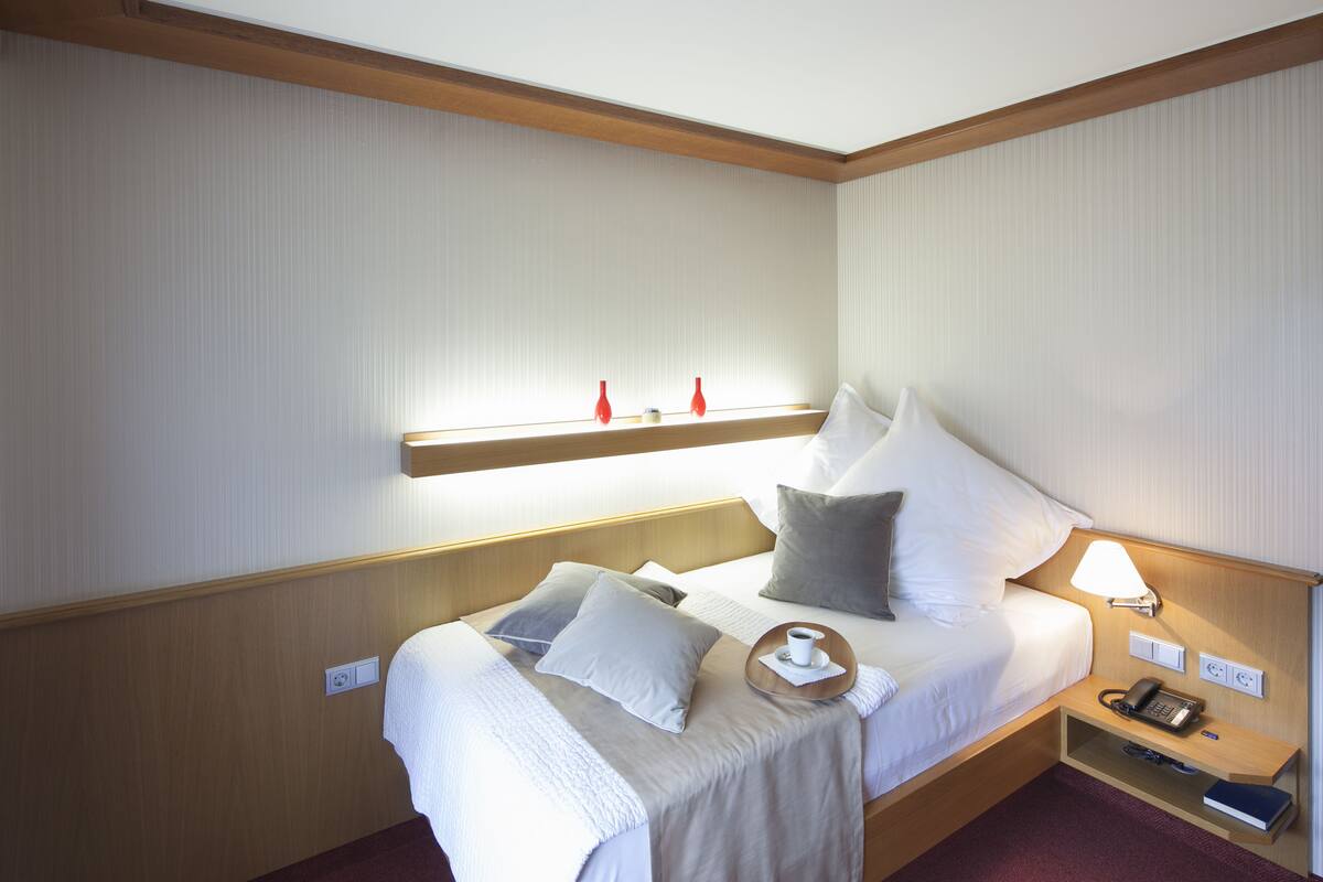 A hotel room with a neatly made bed, bedside table with a lamp, and a small shelf with decorative items.