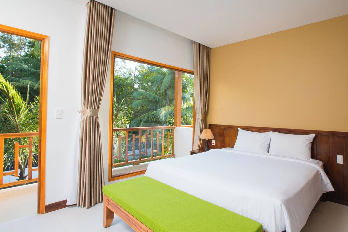A bedroom with a large bed, a wooden headboard, a nightstand, a lamp, and a view of a balcony with curtains.