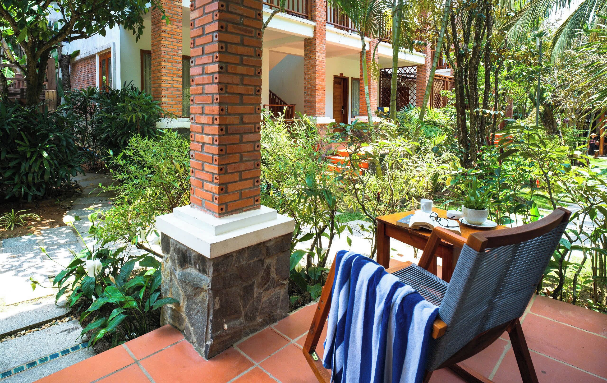 A patio area with a brick pillar, a wooden chair with a towel, a table with a book and a cup, and lush greenery.