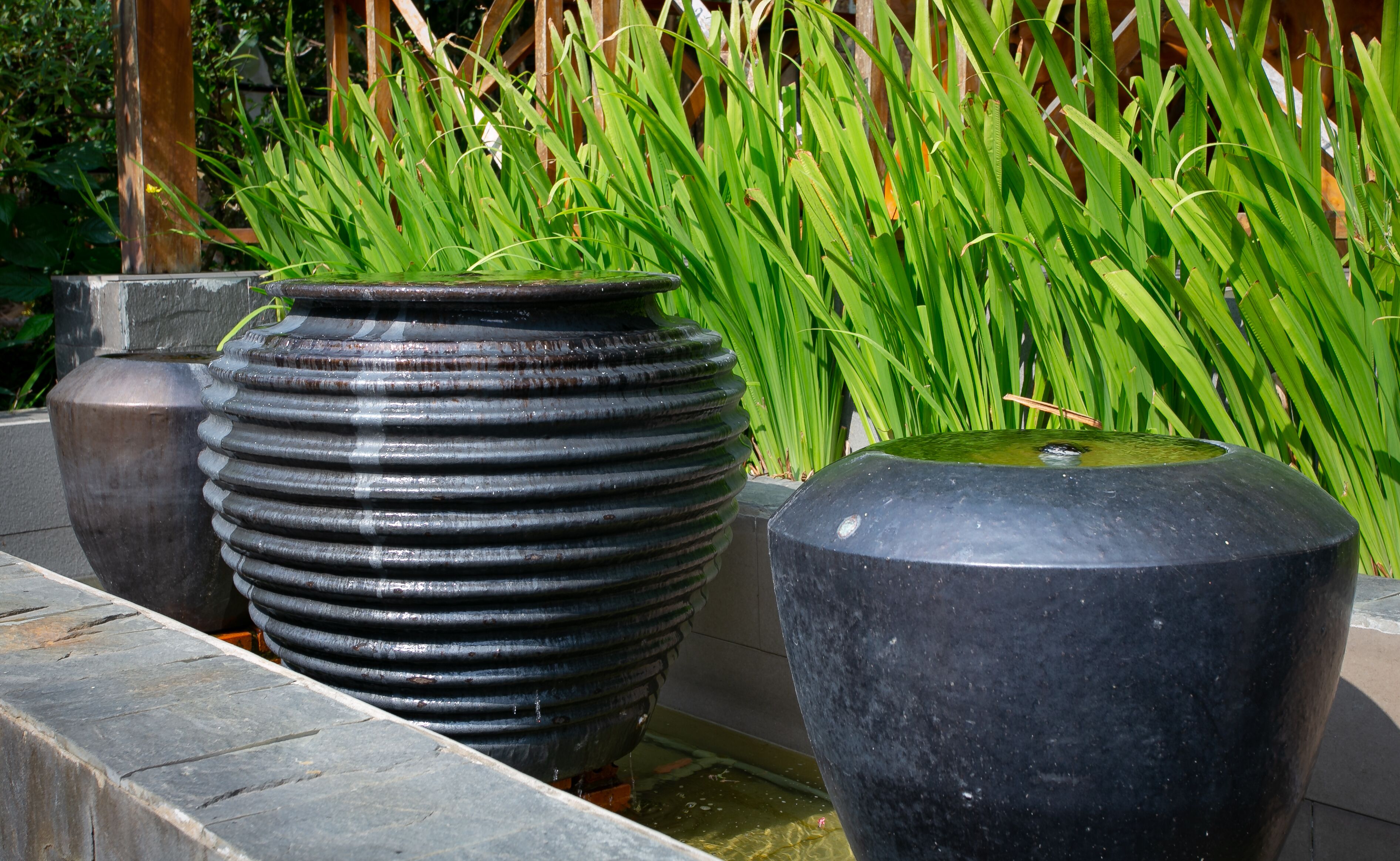 Tres macetas grandes y negras con agua y plantas verdes en un jardín.