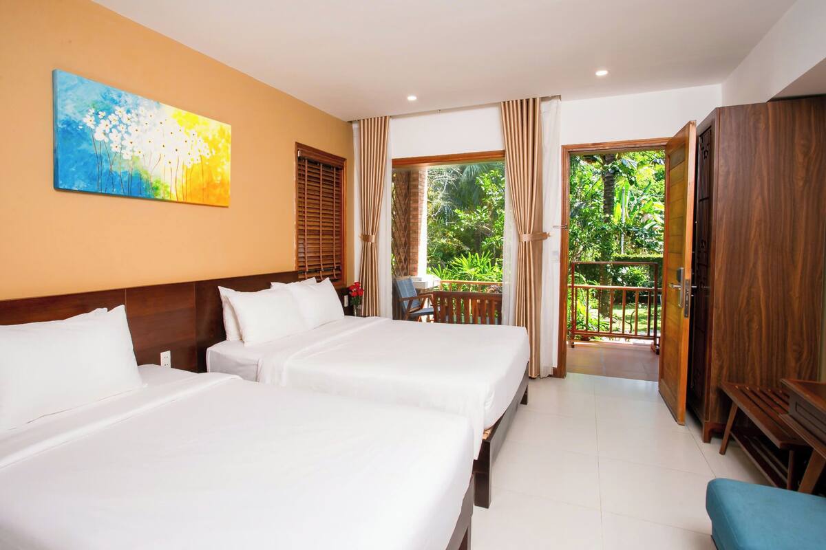 A hotel room with two beds, a painting on the wall, and a balcony with a view of greenery.