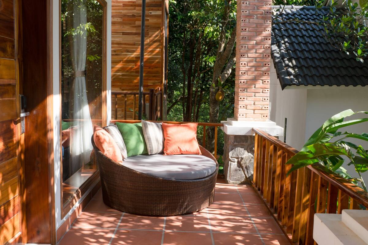 A wicker sofa with cushions on a tiled balcony with a wooden railing and a view of trees.
