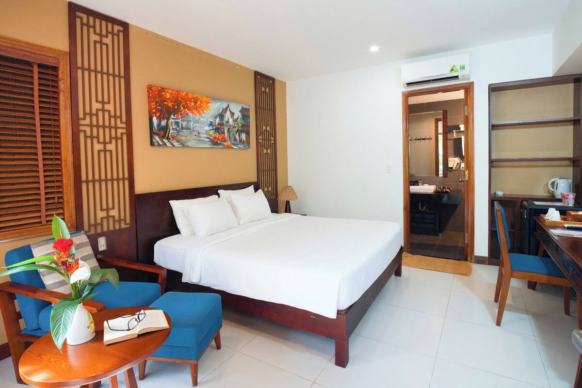 A hotel room with a large bed, a desk, a chair, a small table with a vase and book, and a painting on the wall.