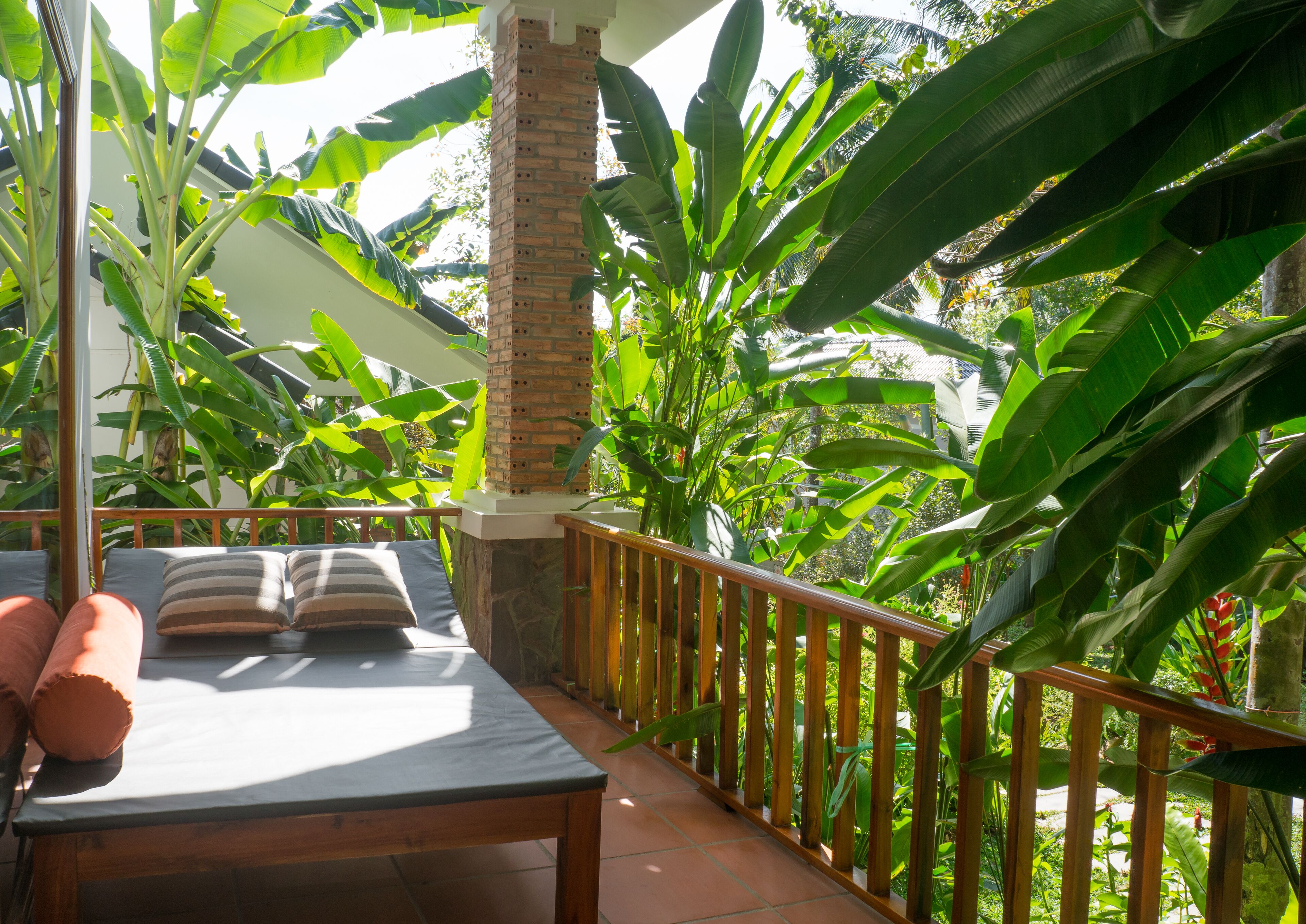 A balcony with a wooden table and two cushioned chairs, surrounded by lush green foliage and a brick pillar.