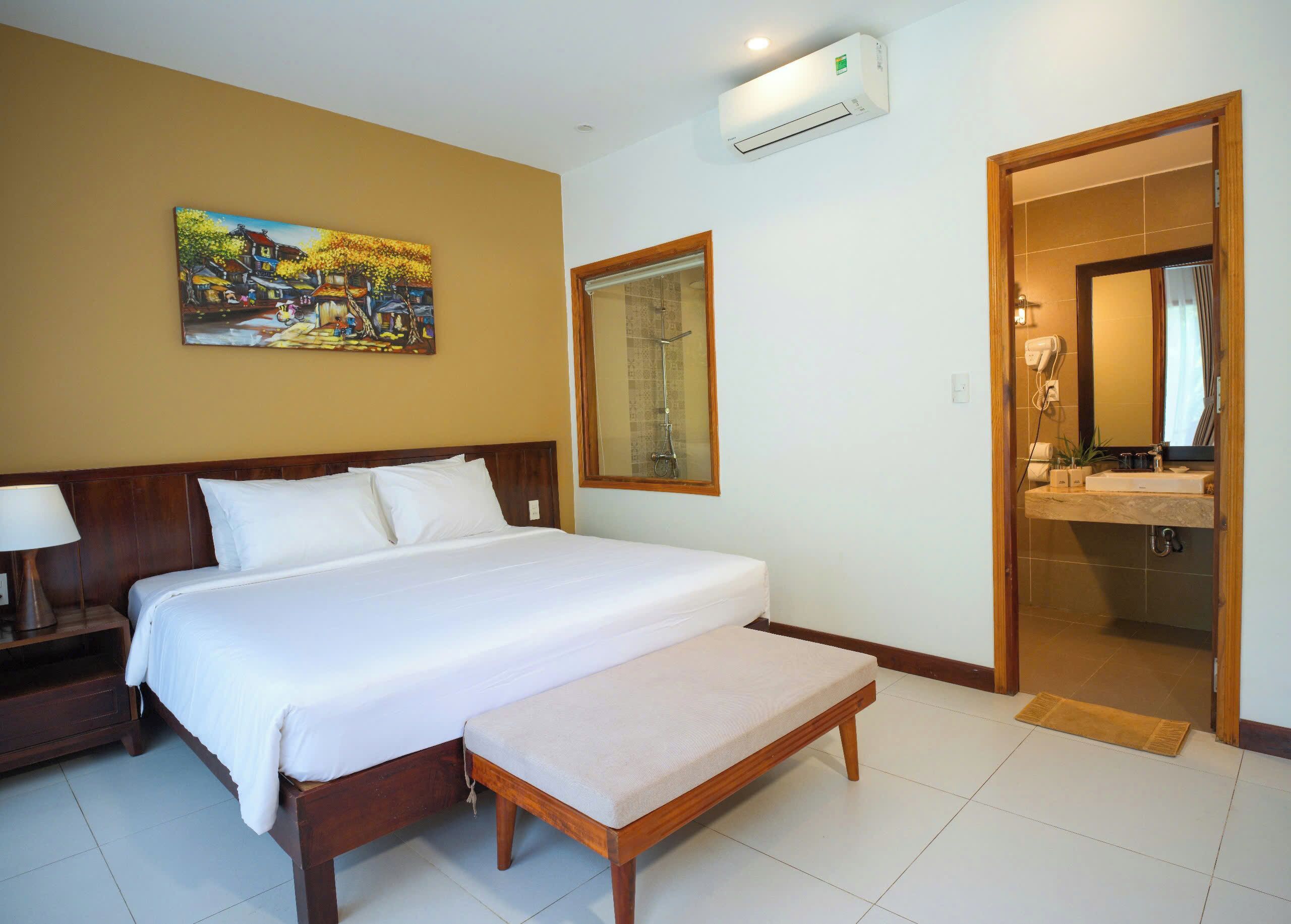 A hotel room with a large bed, a bench, a painting, and a bathroom visible through an open door.