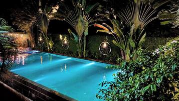 En natlig swimmingpool omgivet af frodig tropisk vegetation og dekorativ belysning.