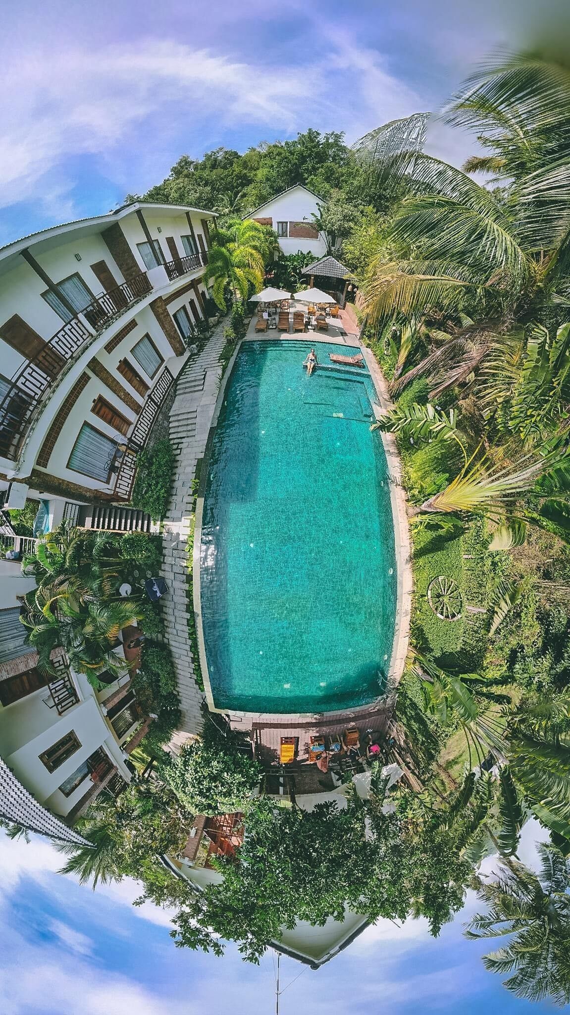 An aerial view of a resort with a central swimming pool surrounded by lush greenery and palm trees.