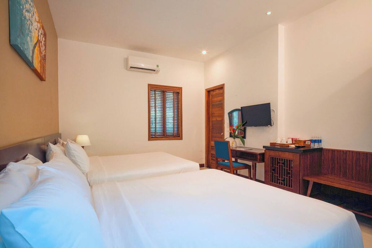 A hotel room with two beds, a desk with a chair, a TV, and an air conditioning unit.