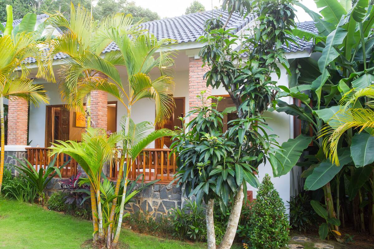 A house with a brick exterior, a wooden porch, and lush tropical vegetation.