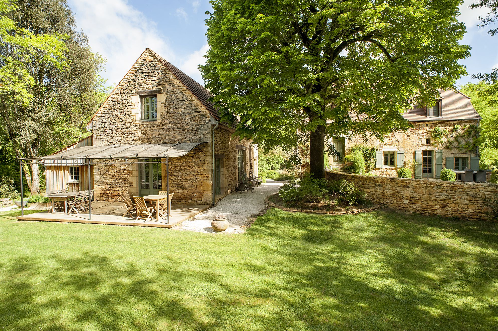Une maison en pierre avec un toit en pente, une terrasse avec des meubles d’extérieur et un grand arbre au premier plan.