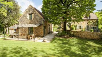 Une maison en pierre avec un toit en pente, une terrasse avec des meubles d’extérieur et un grand arbre au premier plan.
