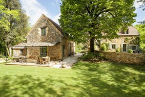 Ein Haus aus Stein mit einem geneigten Dach, einer Terrasse mit Gartenmöbeln und einem großen Baum im Vordergrund.