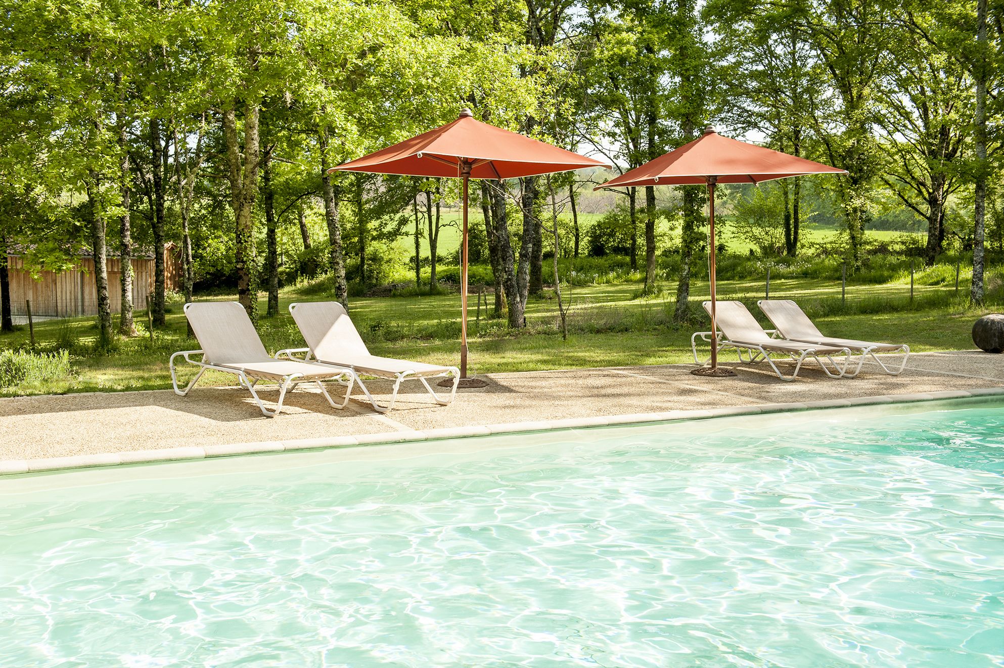 Un espace aménagé au bord de la piscine, avec deux chaises longues et deux parasols rouges, entouré d’arbres et d’une pelouse.