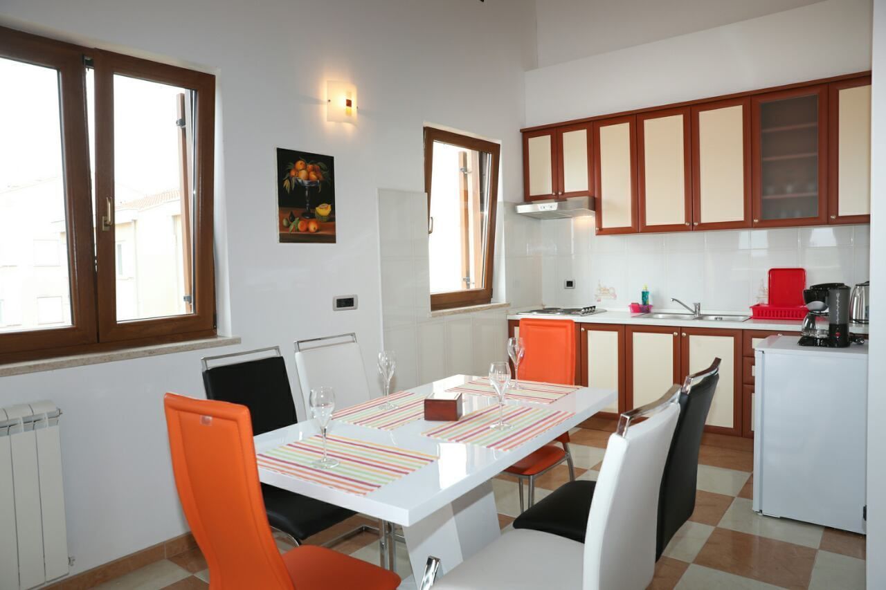 A modern kitchen with white cabinets, a dining area with a white table and orange chairs, and a window with a view of buildings outside.