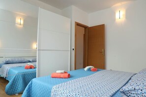 Une chambre d’hôtel avec deux lits, chacun recouvert d’une courtepointe bleue matelassée et agrémenté d’un oreiller blanc. Des serviettes sont posées sur les lits, et la chambre est dotée d’une porte en bois et d’un mur blanc avec un luminaire.