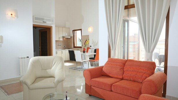 Ein modernes Wohnzimmer mit einem Glastisch, einem beigefarbenen Sofa und einem orangenen Sofa. Im Hintergrund ist eine Küche mit weißen Schränken und einem Essbereich zu sehen.
