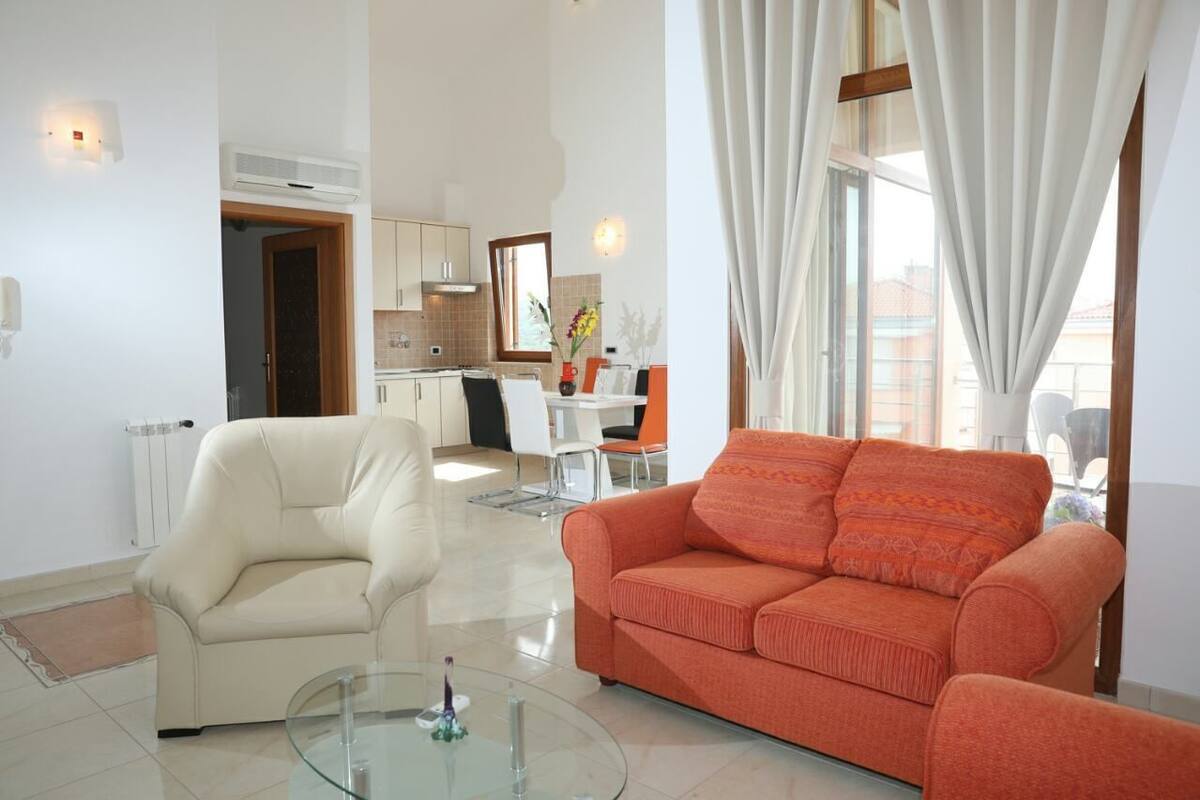 A modern living room with a glass coffee table, a beige sofa, and an orange sofa. There is a kitchen in the background with white cabinets and a dining area.